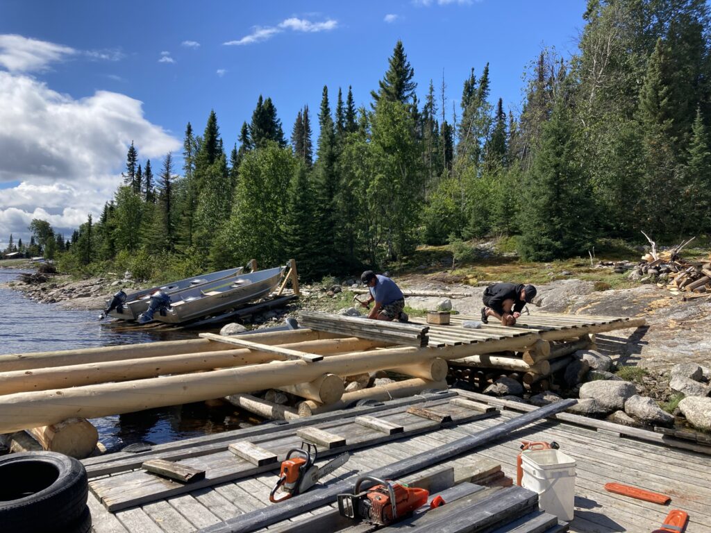 Two people are building a wooden dock by a forested lakeshore. A boat is in the water, and tools are scattered on the platform.