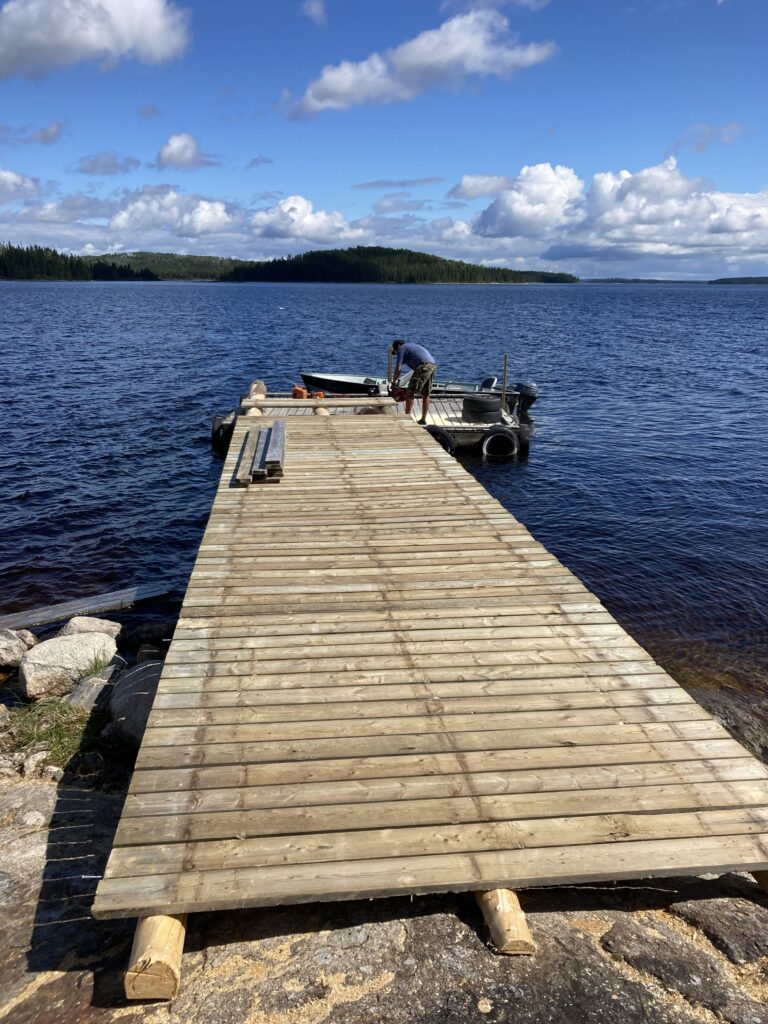 Person standing on a wooden dock extending into a lake, next to a small boat, with a forested shoreline in the background.