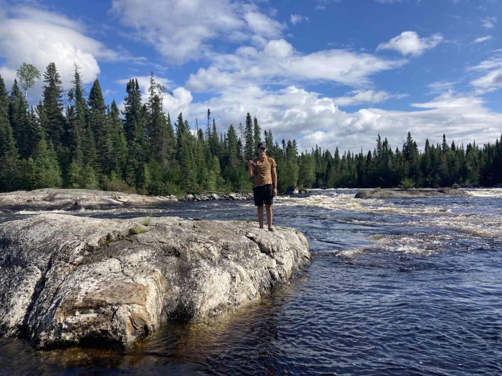 Person standing on a rock in a river, surrounded by trees, with a bright blue sky and clouds above.