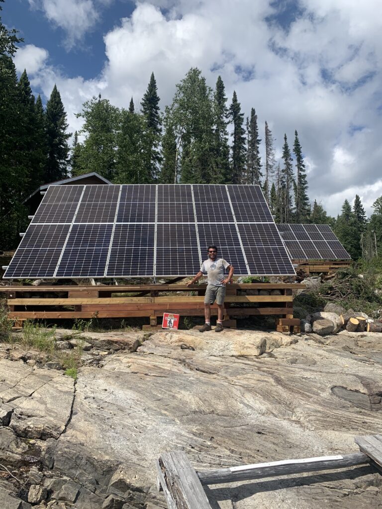 A person stands near a large solar panel array on wooden supports, with trees in the background and a rocky foreground.