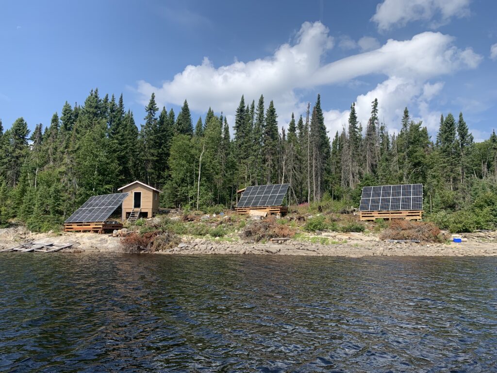 Three small wooden cabins with solar panels are situated by a lake, surrounded by a dense forest under a partly cloudy sky.