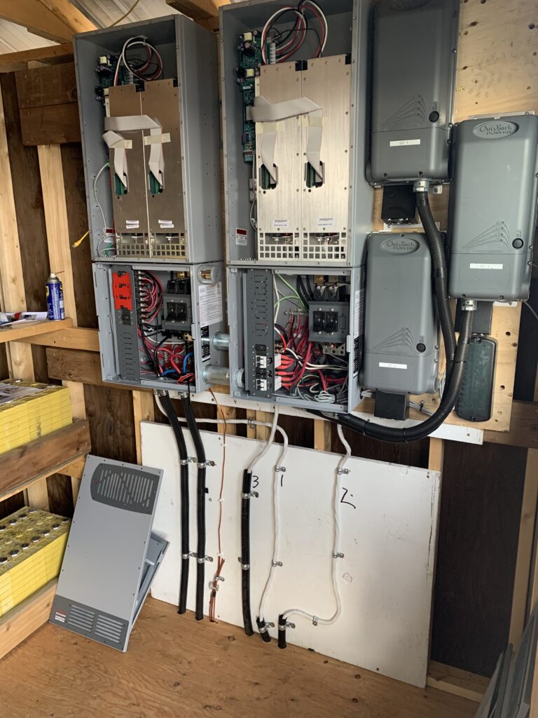 Electrical panel with multiple circuit breakers, wiring, and conduits in a wooden enclosure. Nearby, a detached panel cover.