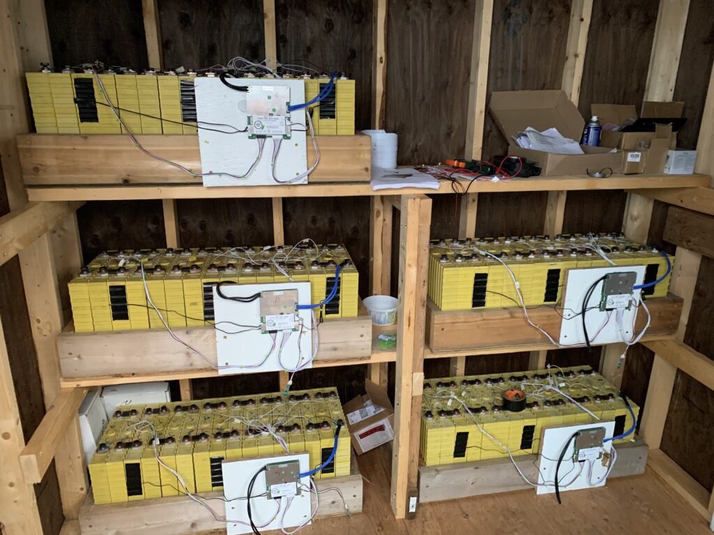 Shelves with yellow battery packs and wiring in a wooden room, alongside tools and miscellaneous items.