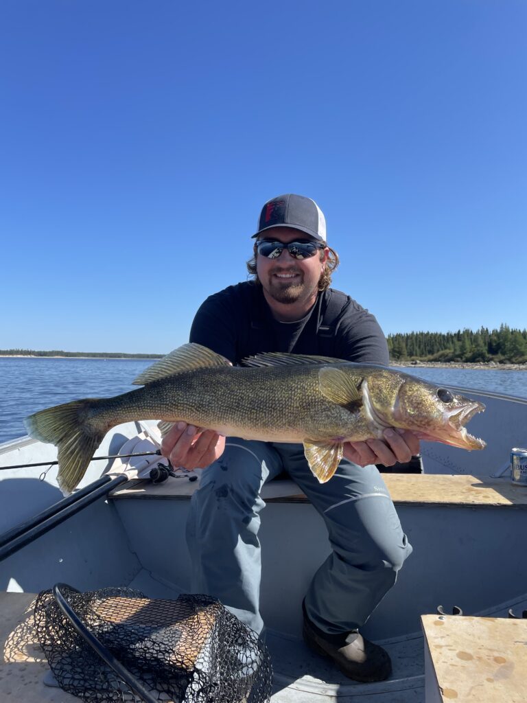 Person on a boat holding a large fish, with a calm lake and forested shoreline in the background under a clear blue sky.