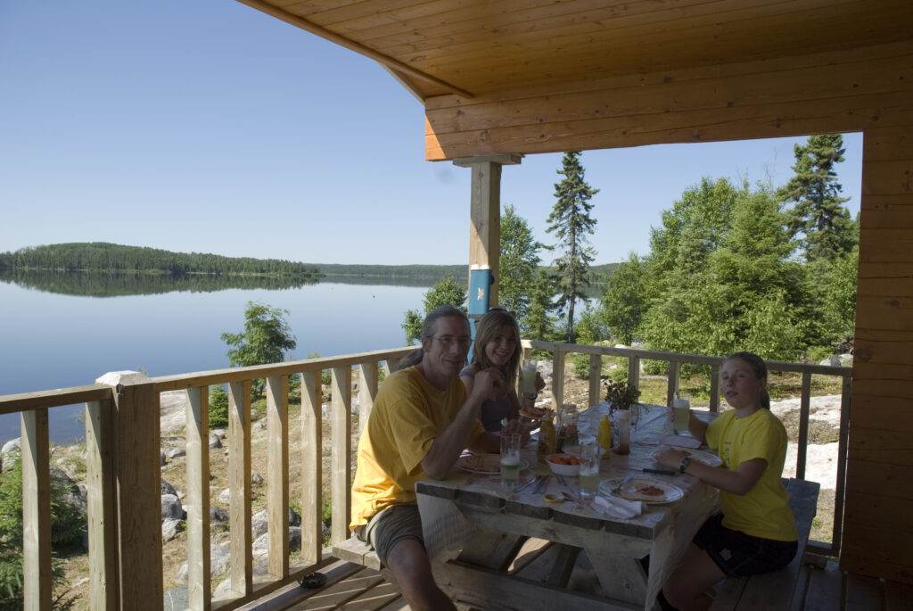 Three people in yellow shirts sit at a wooden table on a porch with trees and a lake view. Plates and food are on the table.