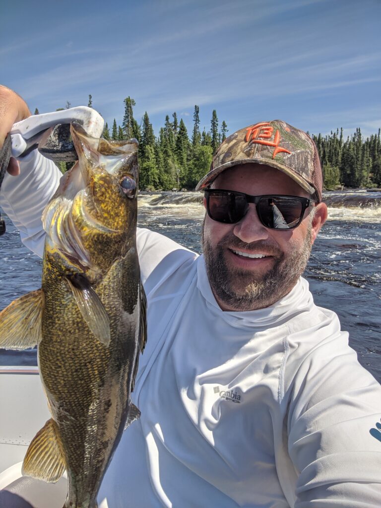 Man in sunglasses and cap holds a large fish on a boat, with a forest and river in the background.