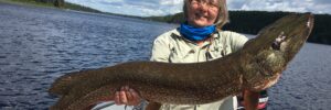 A person in a boat holding a large northern pike against a backdrop of a lake and forest under a partly cloudy sky.