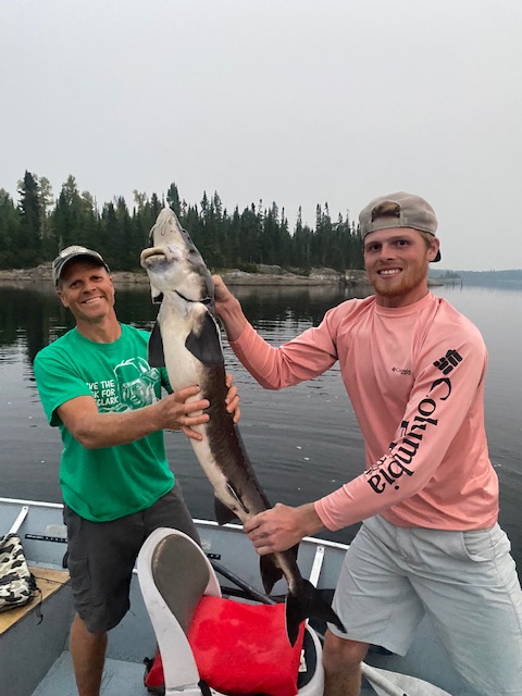 Two men on a boat holding a large fish with a lake and trees in the background.