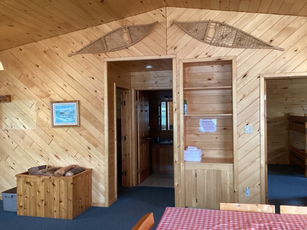 Wood-paneled cabin interior with a doorway, a small sink area, snowshoes on the wall, and a stack of firewood in a box.